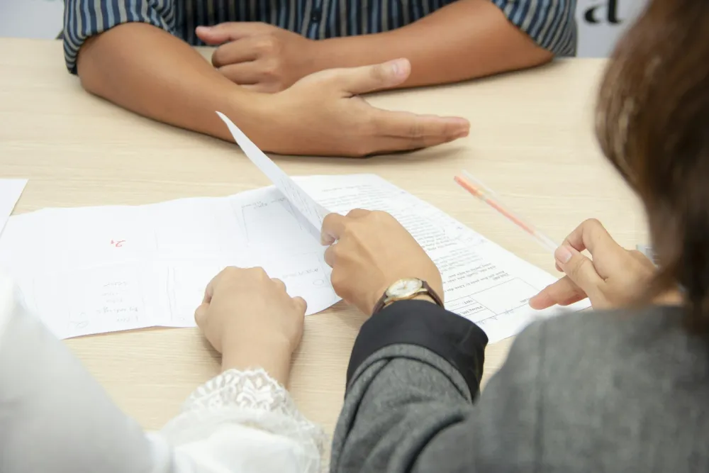 Two people seated across a light wooden table reviewing printed documents, one gesturing toward the page as they talk.